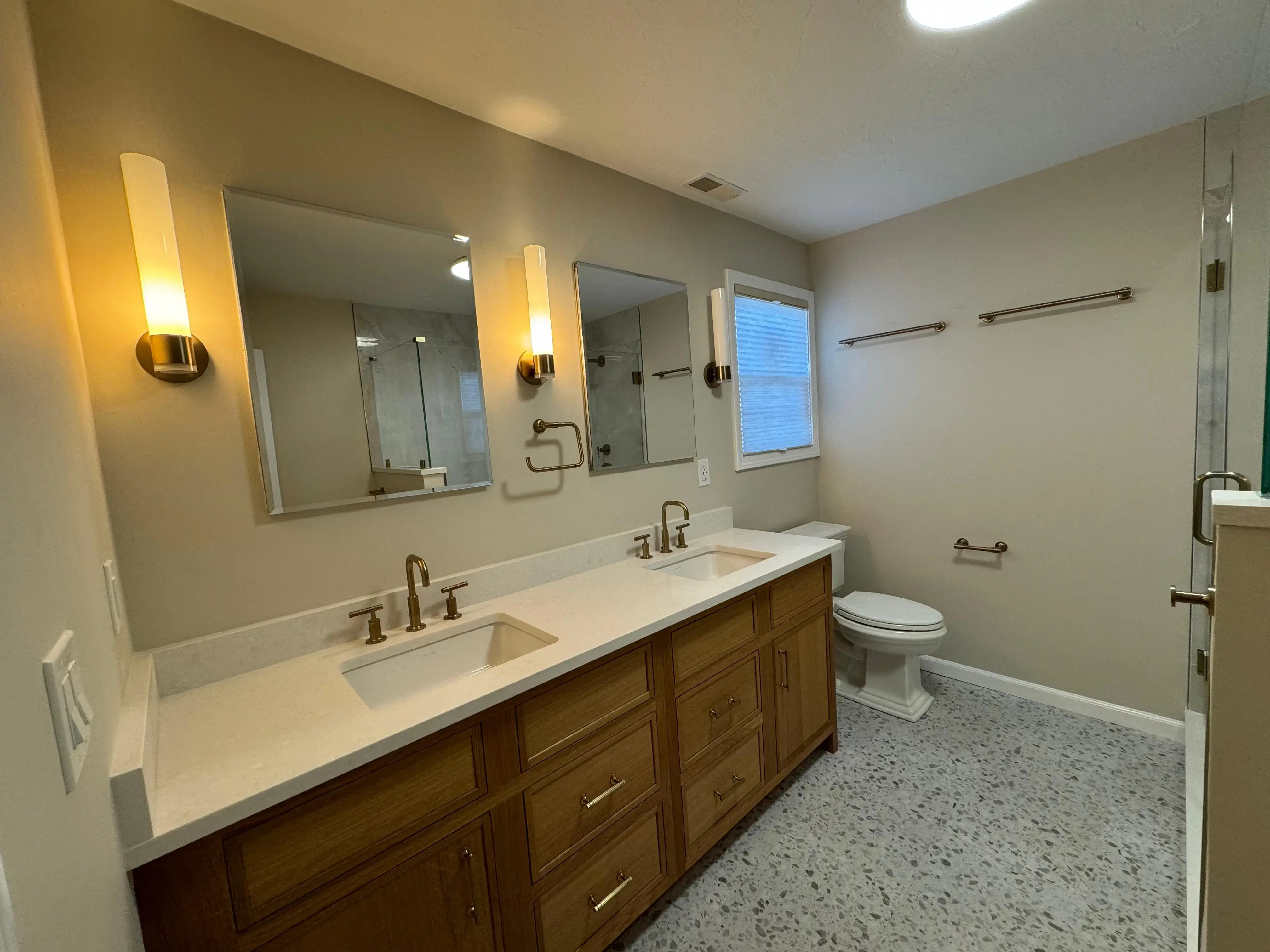 Primary bathroom showing double vanity with wood cabinets, gold faucets, and terrazzo tile floor.