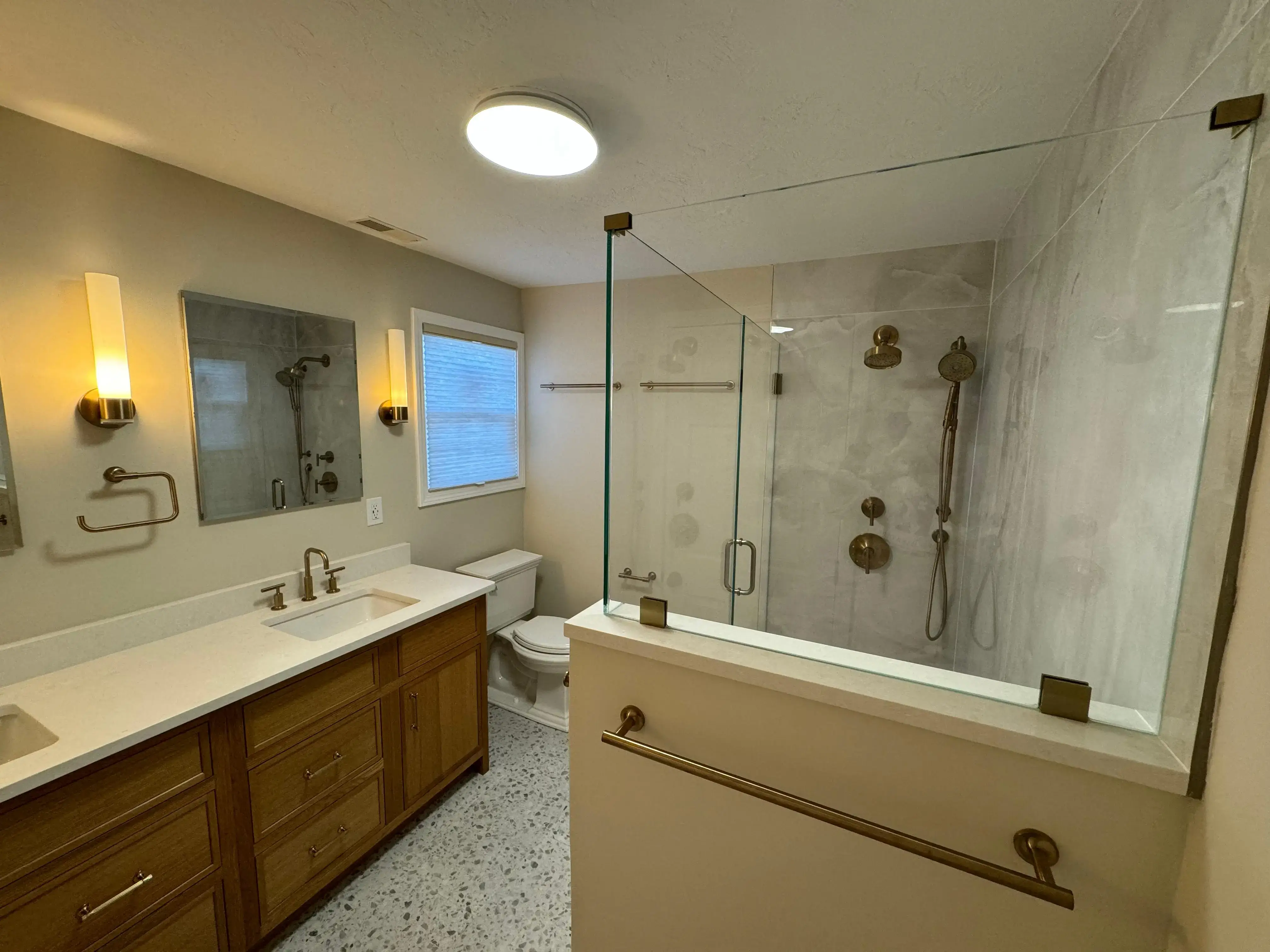 Bathroom remodel with wood double vanity, terrazzo tiled floor, and custom glass shower enclosure.